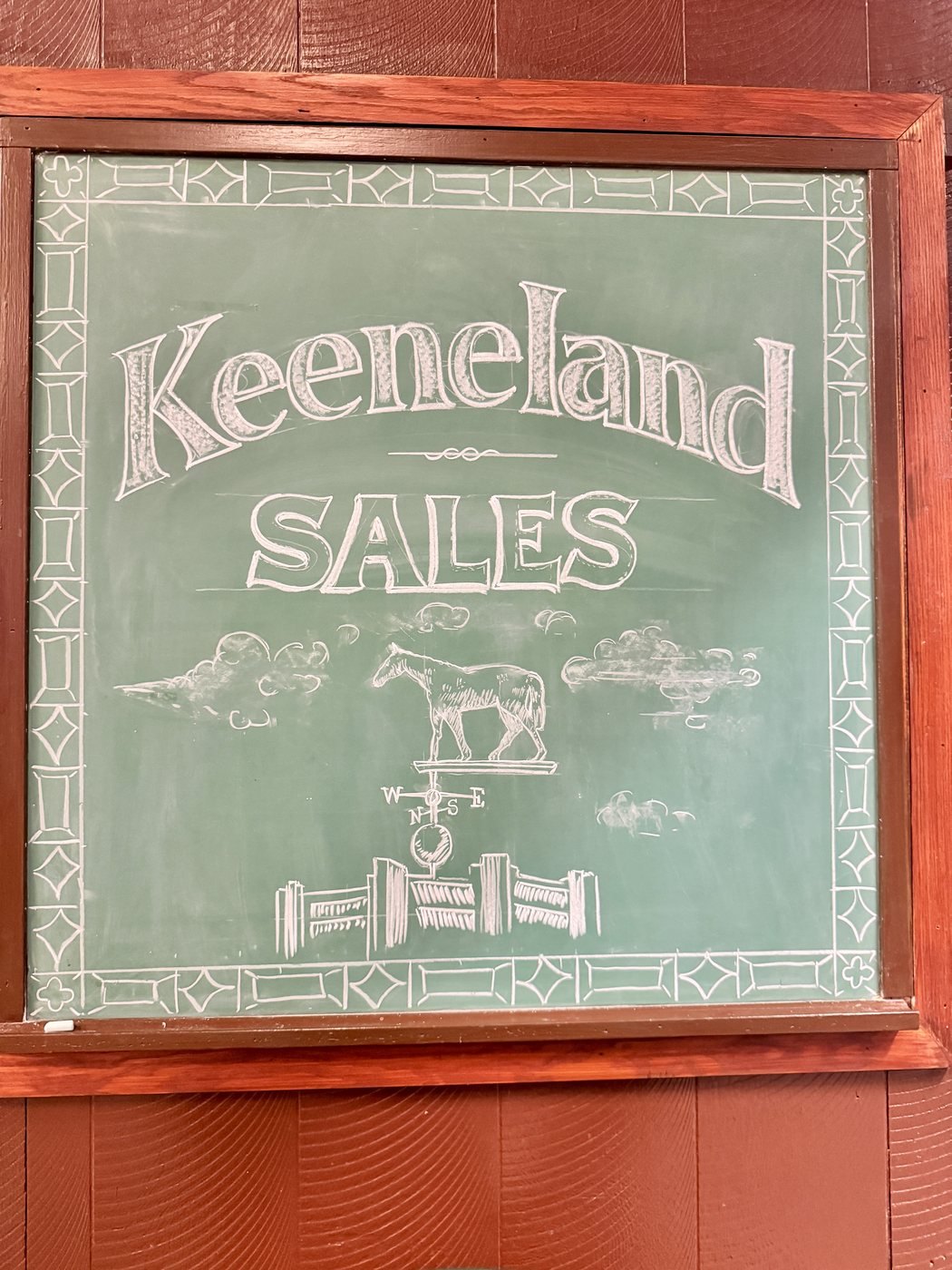 A framed chalkboard sign reading Keeneland Sales, hand lettered in white chalk on green, with a horse weathervane drawn above a plank fence.