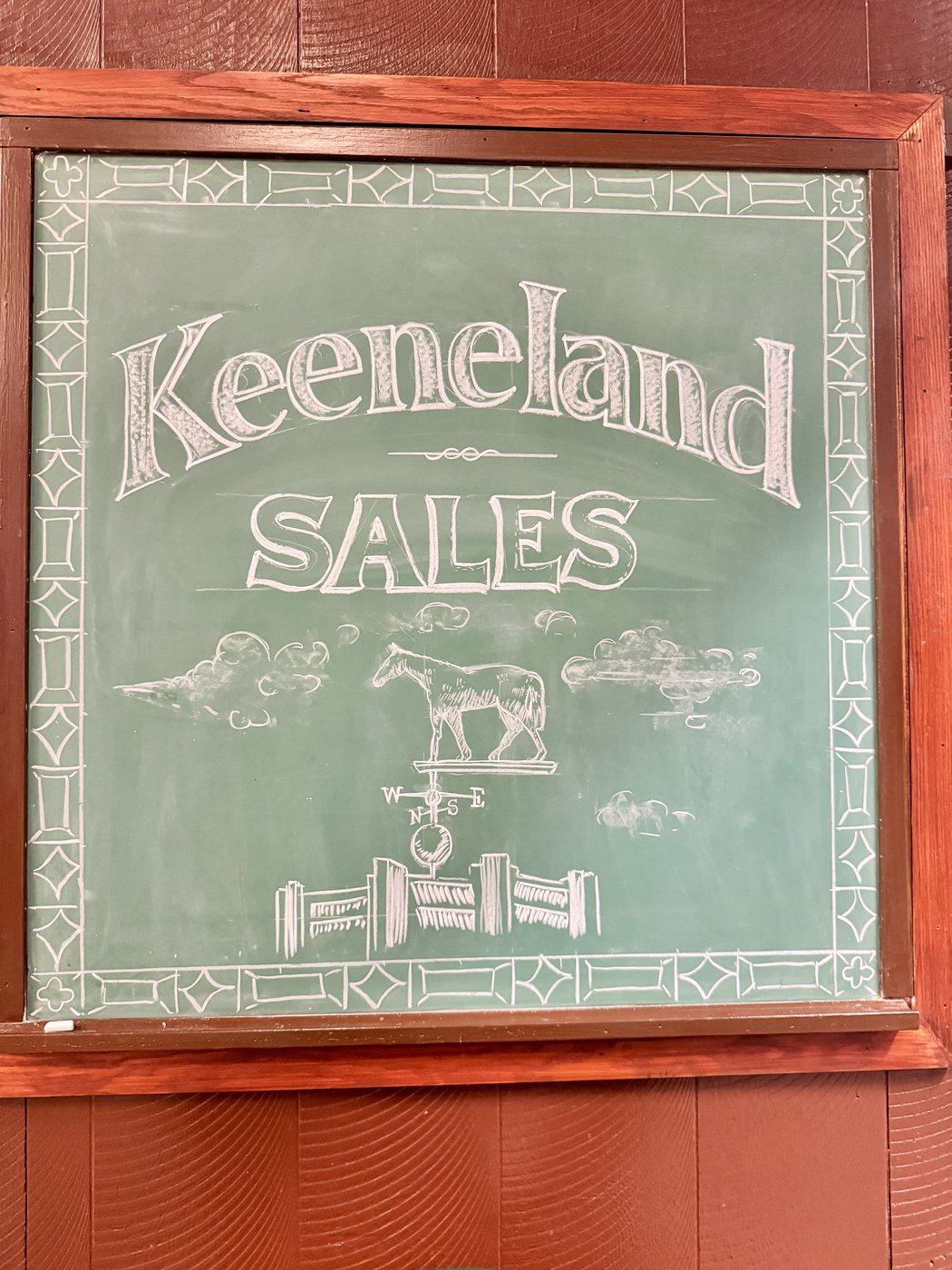 A chalkboard sign lettered by hand reading Keeneland Sales, with a drawing of a weathervane horse above a fence.